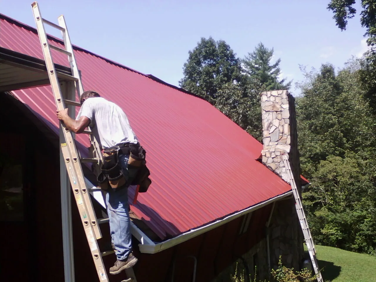 Roofer on a ladder beside a red metal roof and stone chimney — Soffit & Fascia Repair by Roofing Brothers in Absarokee